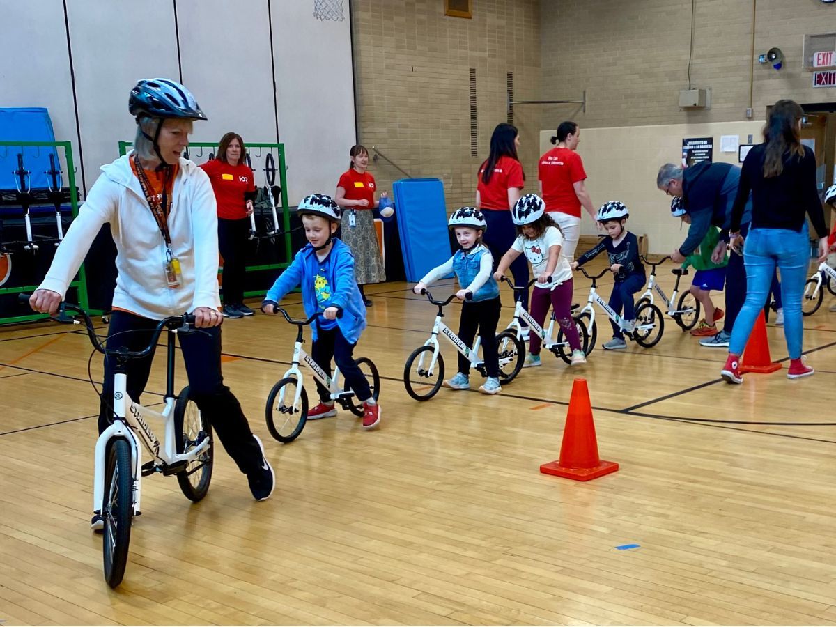 A teacher on a balance bike leads school children on balance bikes around an orange cone.