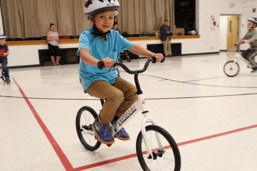 A child with a helmet on rides on a balance bike in a gymnasium.