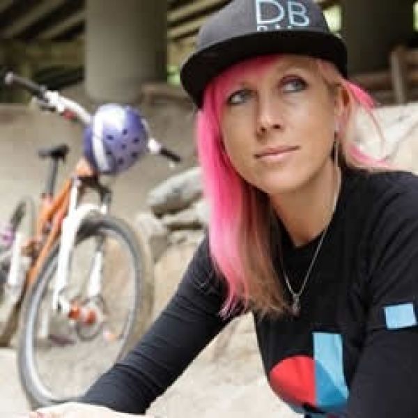 A portrait of All Kids Bike ambassador Kat Sweet with dyed pink hair and a trucker hat and bikes in the background