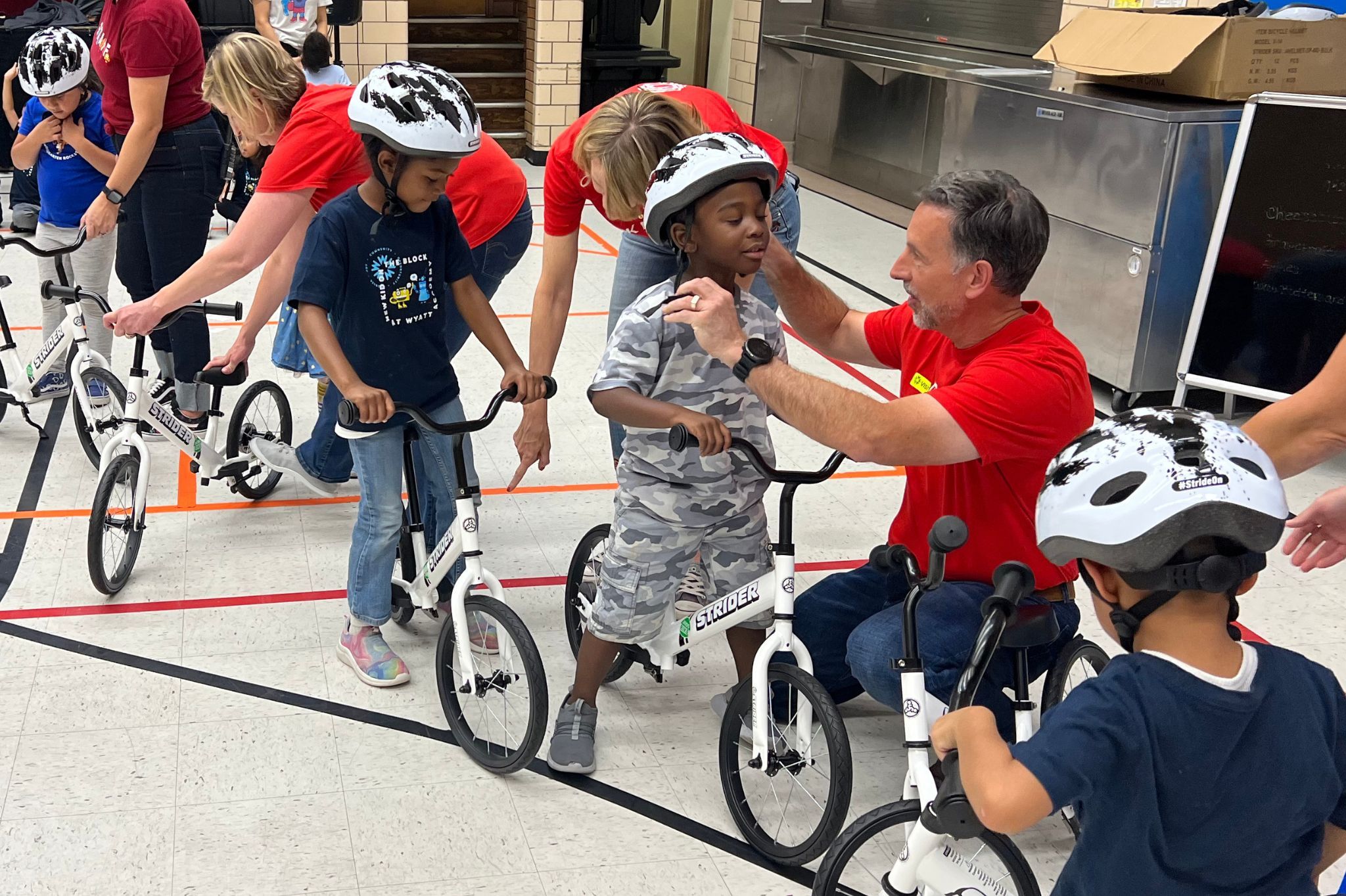 Adults helping children to put on helmets for biking lesson in kindergarten PE gym class