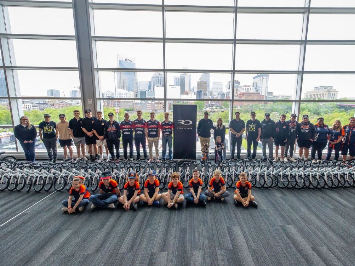 A group of adults and children pose behind Strider balance bikes, children sit in front of the bikes.