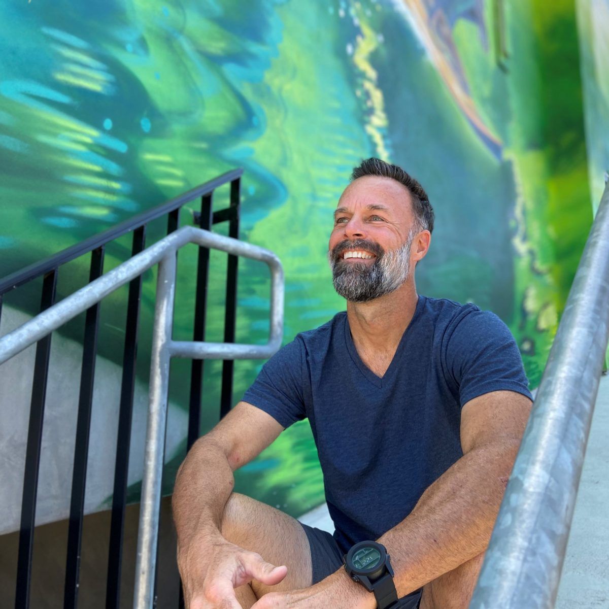 Greg Lenac sits on a stairway next to a blue and green mural