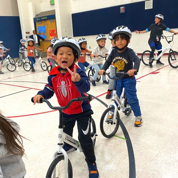 A smiling child in a red Spiderman helmet poses with a blue Strider 14x balance bike