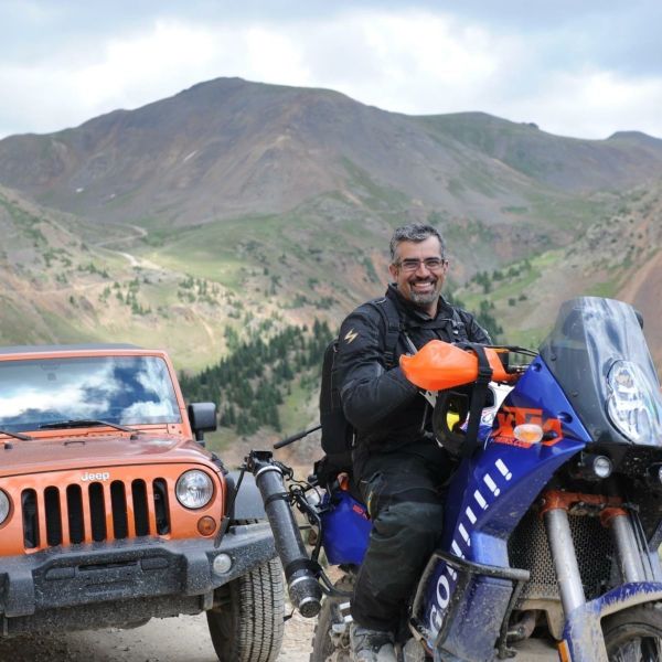 All Kids Bike ambassador Robert Pandya on a blue motocycle with an orange jeep and a mountainscape in the background
