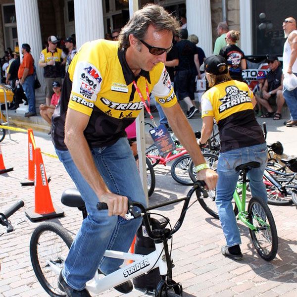 Ryan McFarland, wearing a yellow and black Strider bike jersey and blue jeans, demonstrates how to ride a balance bike during an outdoor event