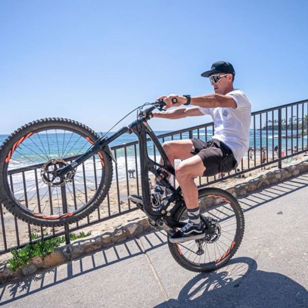 All Kids Bike ambassador Brian Lopez popping a wheelie on a mountain bike with a beach in the background