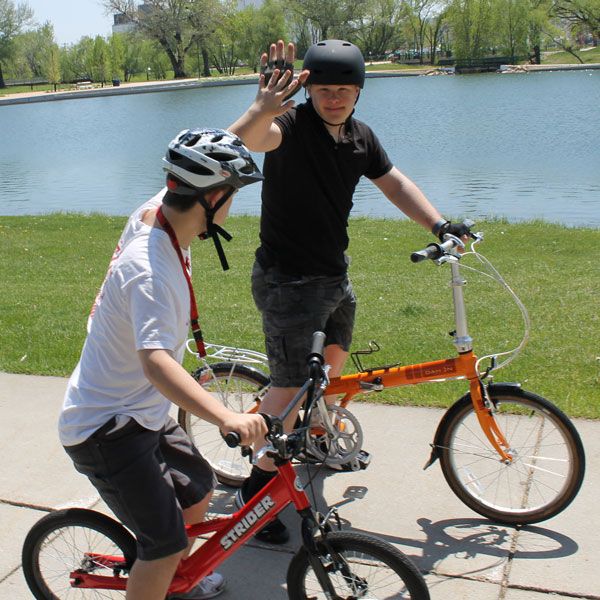 Two bikers, one on an aroange bike and one on a red balance bike, high five on a lakeside path.